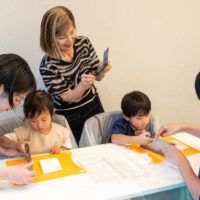 子ども2人が大人に見守られながら、包丁でうどんを切っている。Two children cutting udon noodles with knives while adults look on. 出張手打ちうどん作り体験 2026年8月8日 20260808-4.jpg – イリコスキー製麺所
